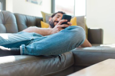 Genç sakallı adam akıllı telefonuyla kanepede dinleniyor.