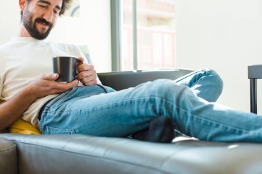 Genç sakallı adam elinde kahve fincanıyla kanepede dinleniyor.