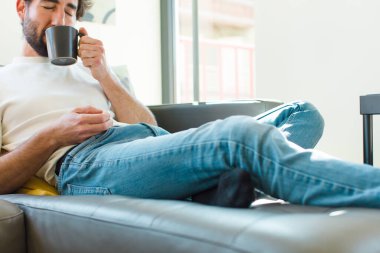 Genç sakallı adam elinde kahve fincanıyla kanepede dinleniyor.