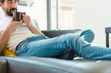 Genç sakallı adam elinde kahve fincanıyla kanepede dinleniyor.