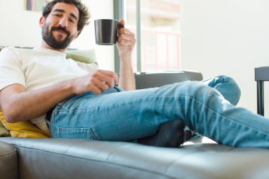 Genç sakallı adam elinde kahve fincanıyla kanepede dinleniyor.