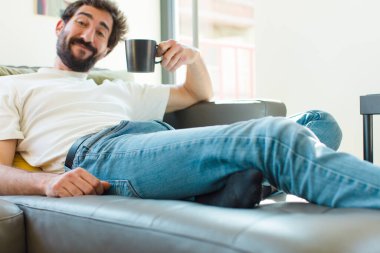 Genç sakallı adam elinde kahve fincanıyla kanepede dinleniyor.