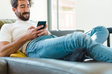Genç sakallı adam akıllı bir telefonla kanepede dinleniyor.