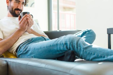 Genç sakallı adam elinde kahve fincanıyla kanepede dinleniyor.