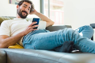 Genç sakallı adam akıllı bir telefonla kanepede dinleniyor.