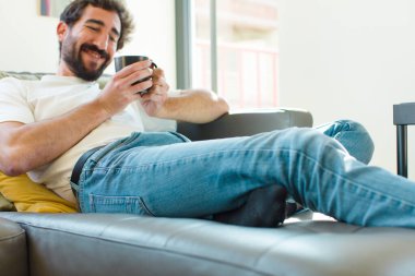 Genç sakallı adam elinde kahve fincanıyla kanepede dinleniyor.