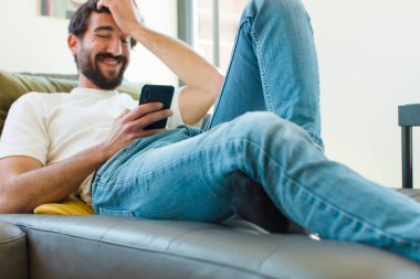 Genç sakallı adam akıllı bir telefonla kanepede dinleniyor.