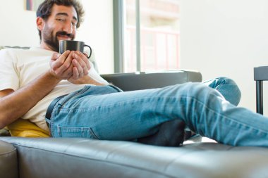 Genç sakallı adam elinde kahve fincanıyla kanepede dinleniyor.