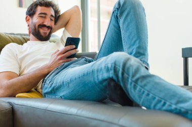 Genç sakallı adam akıllı bir telefonla kanepede dinleniyor.