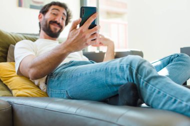Genç sakallı adam akıllı bir telefonla kanepede dinleniyor.