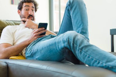 Genç sakallı adam akıllı bir telefonla kanepede dinleniyor.