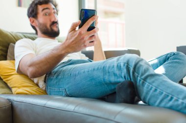 Genç sakallı adam akıllı bir telefonla kanepede dinleniyor.