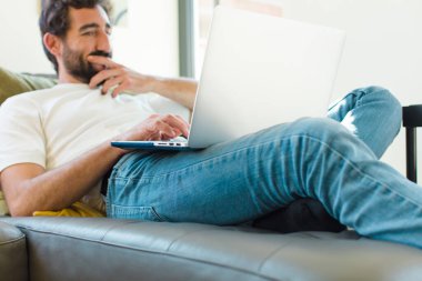 Genç sakallı bir adam dizüstü bilgisayarla kanepede dinleniyor.