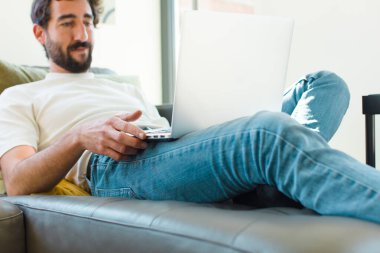 Genç sakallı bir adam dizüstü bilgisayarla kanepede dinleniyor.