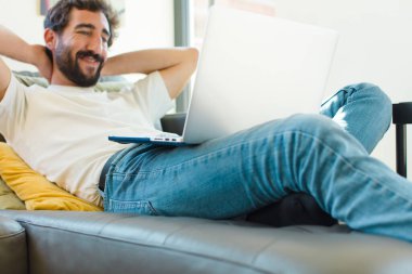 Genç sakallı bir adam dizüstü bilgisayarla kanepede dinleniyor.