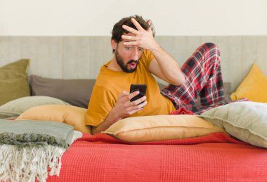 Yatakta cep telefonuyla sakallı genç bir adam.