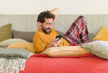 Yatakta cep telefonuyla sakallı genç bir adam.