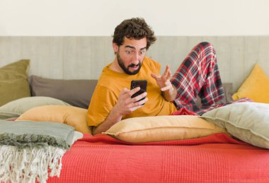 Yatakta cep telefonuyla sakallı genç bir adam.
