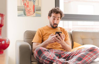 Genç sakallı adam akıllı bir telefonla kanepede oturuyor.