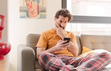 Genç sakallı adam akıllı bir telefonla kanepede oturuyor.