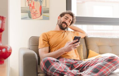 Genç sakallı adam akıllı bir telefonla kanepede oturuyor.
