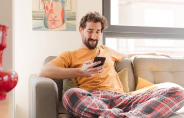 Genç sakallı adam akıllı bir telefonla kanepede oturuyor.