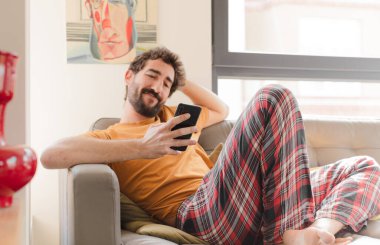 Genç sakallı adam akıllı bir telefonla kanepede oturuyor.