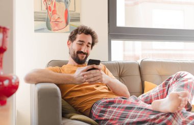 Genç sakallı adam akıllı bir telefonla kanepede oturuyor.