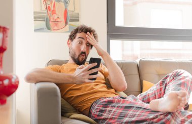 Genç sakallı adam akıllı bir telefonla kanepede oturuyor.