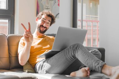 Genç sakallı adam gülümsüyor ve mutlu görünüyor, kaygısız ve pozitif, tek eliyle bir dizüstü bilgisayarla zafer ya da barışı işaret ediyor.