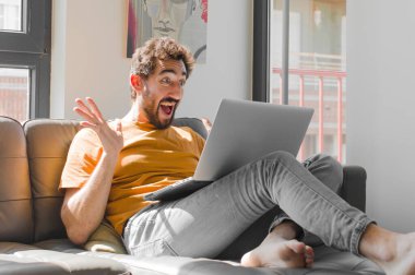 Genç sakallı adam mutlu, heyecanlı, şaşırmış ya da şok olmuş hissediyor. Kanepe dizüstü bilgisayarında inanılmaz bir şeye gülümsüyor ve hayrete düşüyor.