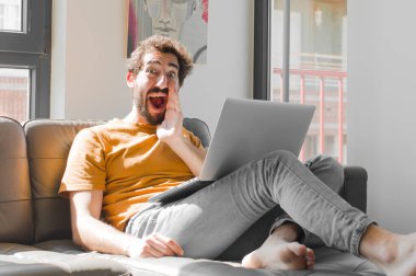 Genç sakallı adam mutlu, heyecanlı ve pozitif hissediyor, elleri ağızda bağırıyor, dizüstü bilgisayarla kanepe konseptinde bağırıyor.