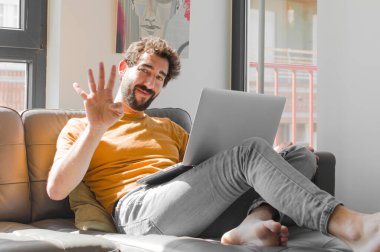 Genç sakallı adam gülümsüyor ve arkadaş canlısı görünüyor. Elleri önde, dizüstü bilgisayarında dizüstü bilgisayarla dört ya da dört numarayı gösteriyor.