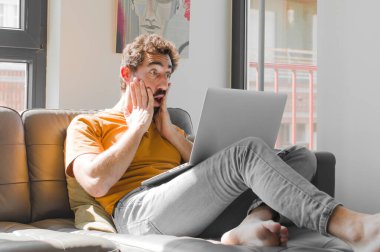 Genç sakallı adam şok olmuş ve korkmuş hissediyor, ağzı açık ve elleri yanaklarında, dizüstü bilgisayarlı kanepe konseptinde.