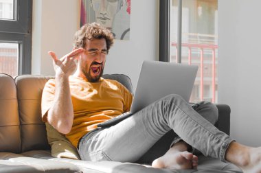 Genç sakallı adam mutsuz ve stresli görünüyor, intihar jesti elle silah işareti yapıyor, kanepe dizüstü bilgisayarındaki bir bilgisayarı işaret ediyor.