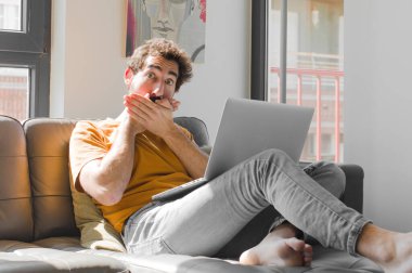 Genç sakallı bir adam elleri ile ağzını kapatıyor, şok olmuş, şaşırmış bir ifadeyle, bir sır saklıyor ya da dizüstü bilgisayarla kanepe konseptinde 