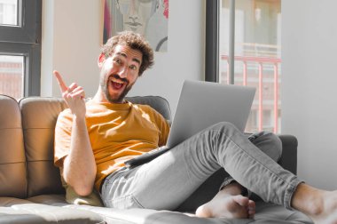 Genç sakallı adam bir fikri fark ettikten sonra mutlu ve heyecanlı bir dahi gibi hissediyor, neşeli bir şekilde parmağını kaldırıyor, eureka! Dizüstü bilgisayarlı kanepe konsepti.