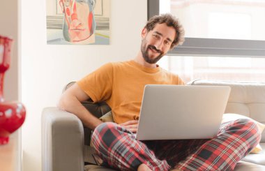 Genç sakallı adam mutlu bir şekilde gülümsüyor, eli kalçasında ve kendinden emin, pozitif, gururlu ve arkadaş canlısı ve dizüstü bilgisayarla oturuyor.