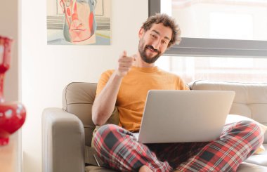 Genç sakallı adam kameraya tatmin olmuş, kendinden emin, dostane bir gülümsemeyle bakıyor, seni seçiyor ve dizüstü bilgisayarla oturuyor.