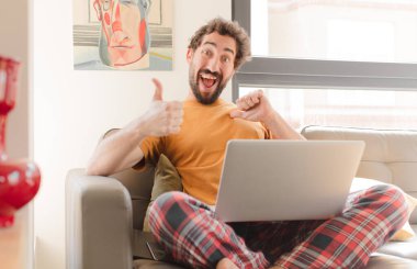 Genç sakallı adam gururlu, kaygısız, kendinden emin ve mutlu hissediyor. Başparmaklarıyla gülümsüyor ve dizüstü bilgisayarla oturuyor.