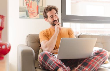 Genç sakallı bir adam mutlu, kendinden emin bir ifadeyle gülümsüyor. Elleri çenesinde, merak ediyor, yan tarafa bakıyor ve dizüstü bilgisayarla oturuyor.