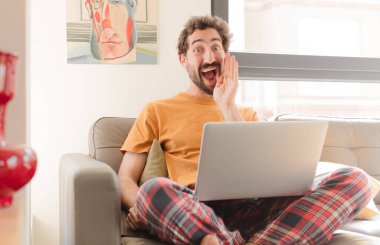 Genç sakallı adam mutlu, heyecanlı ve pozitif hissediyor, elleri ağızının yanında bağırıyor, bağırıyor ve dizüstü bilgisayarla oturuyor.
