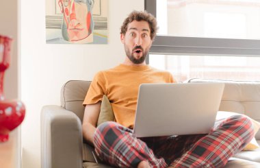 Genç sakallı adam şok olmuş ya da şaşırmış görünüyor, ağzı açık bakıyor ve vay canına diyor ve dizüstü bilgisayarla oturuyor.