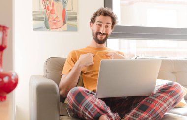 Genç sakallı adam mutlu, şaşırmış ve gururlu hissediyor. Heyecanlı, şaşkın bakışlarıyla ve dizüstü bilgisayarla oturuyor.