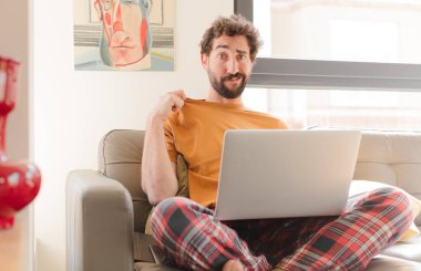 Genç sakallı bir adam stresli, endişeli, yorgun ve hayal kırıklığına uğramış, gömlek yakalı, sorunlu görünüyor ve dizüstü bilgisayarla oturuyor.