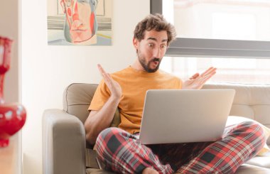 Genç sakallı adam, ağzı açık ve şaşkın, şok olmuş ve hayretler içinde inanılmaz bir sürprizle ve dizüstü bilgisayarla oturuyor.