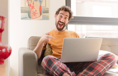 Genç sakallı adam heyecanlı ve şaşırmış görünüyor. Yana ve yukarıya bakıyor ve dizüstü bilgisayarla oturuyor.