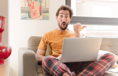 Genç sakallı adam inançsızlıktan şaşkın şaşkın bakıyor, yan taraftaki objeyi işaret ediyor ve diyor ki vay be, inanılmaz ve dizüstü bilgisayarla oturuyor.