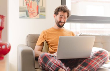 Genç sakallı adam üzgün ve mızmız, mutsuz bir bakışla ağlıyor, olumsuz ve hayal kırıklığına uğramış bir tavırla ve dizüstü bilgisayarla oturuyor.