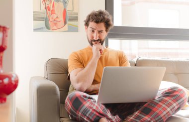 Genç sakallı adam düşünüyor, kuşkulu ve şaşkın hissediyor, farklı seçeneklerle, hangi kararı vereceğini merak ediyor ve bir dizüstü bilgisayarla oturuyor.